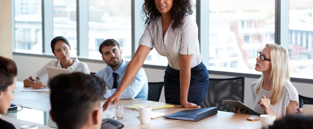 Businesswoman,Stands,To,Address,Meeting,Around,Board,Table